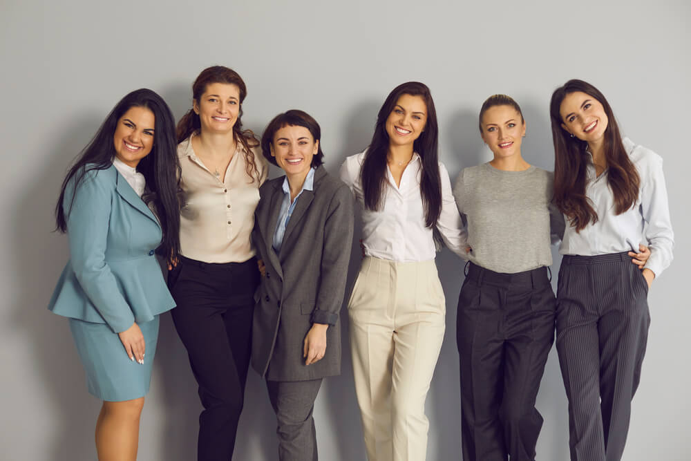 A diverse group of professionally dressed women standing together and posing for the camera against a wall.